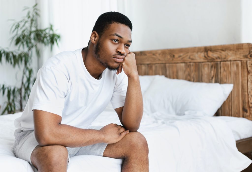 Unhappy Black Guy Sitting On Bed In Bedroom At Home